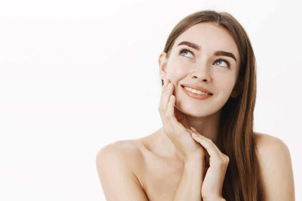 Woman applying cream on face feeling skin soft and tender standing dreamy and delighted with result gazing at upper left corner with sensual smile touching cheek posing naked over grey wall