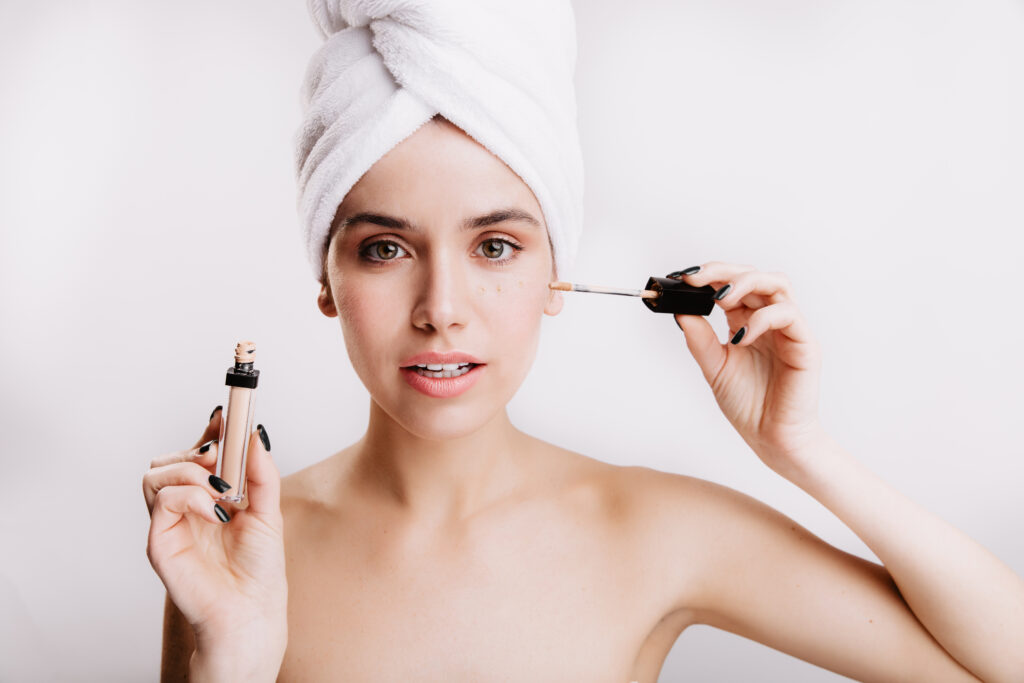 Pretty green-eyed woman looks into camera on white background. Morning routine of model that puts concealer.